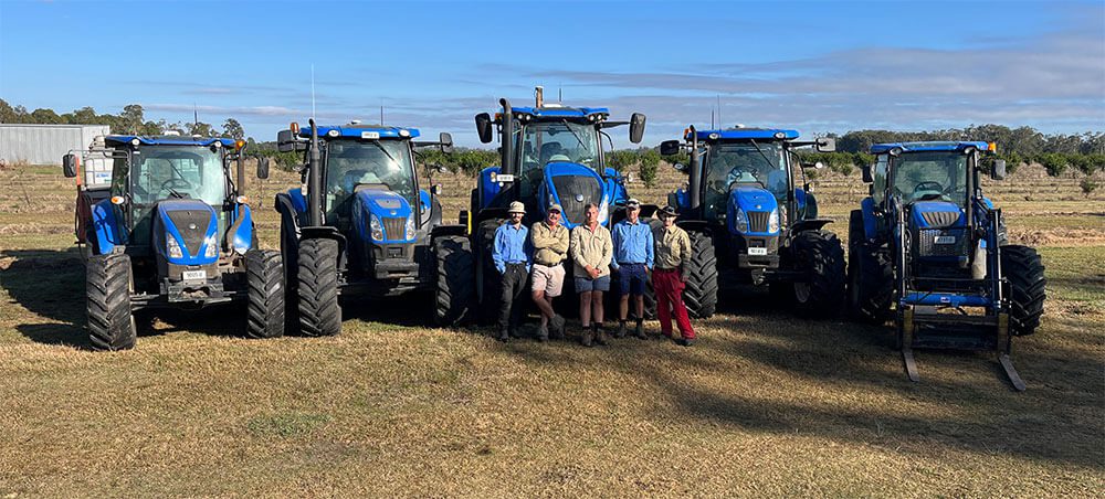 A group of people standing in front of three blue tractors.